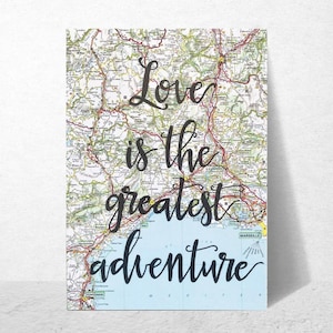 May include: A rectangular print featuring a map with the text "Love is the greatest adventure" in black cursive script. The map has a light blue gradient at the bottom, and the overall color scheme is a mix of greens, browns, and blues.