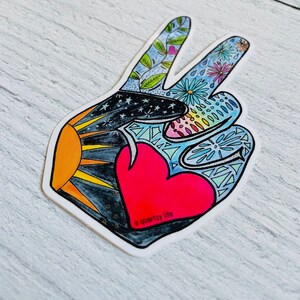 May include: A die-cut sticker in the shape of a peace sign. The design features a red heart, a sun, stars, and floral patterns in various colors. The text "a quartzy life" is printed on the sticker.