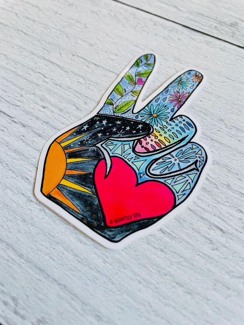 May include: A die-cut sticker in the shape of a peace sign. The design features a red heart, a sun, stars, and floral patterns in various colors. The text "a quartzy life" is printed on the sticker.