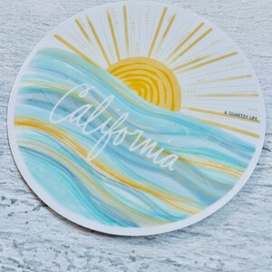May include: A round sticker with a blue and yellow watercolor design of waves and a sun with rays. The text "California" is written in white cursive script and the text "A Quartzy Life" is written in small black letters in the bottom right corner.