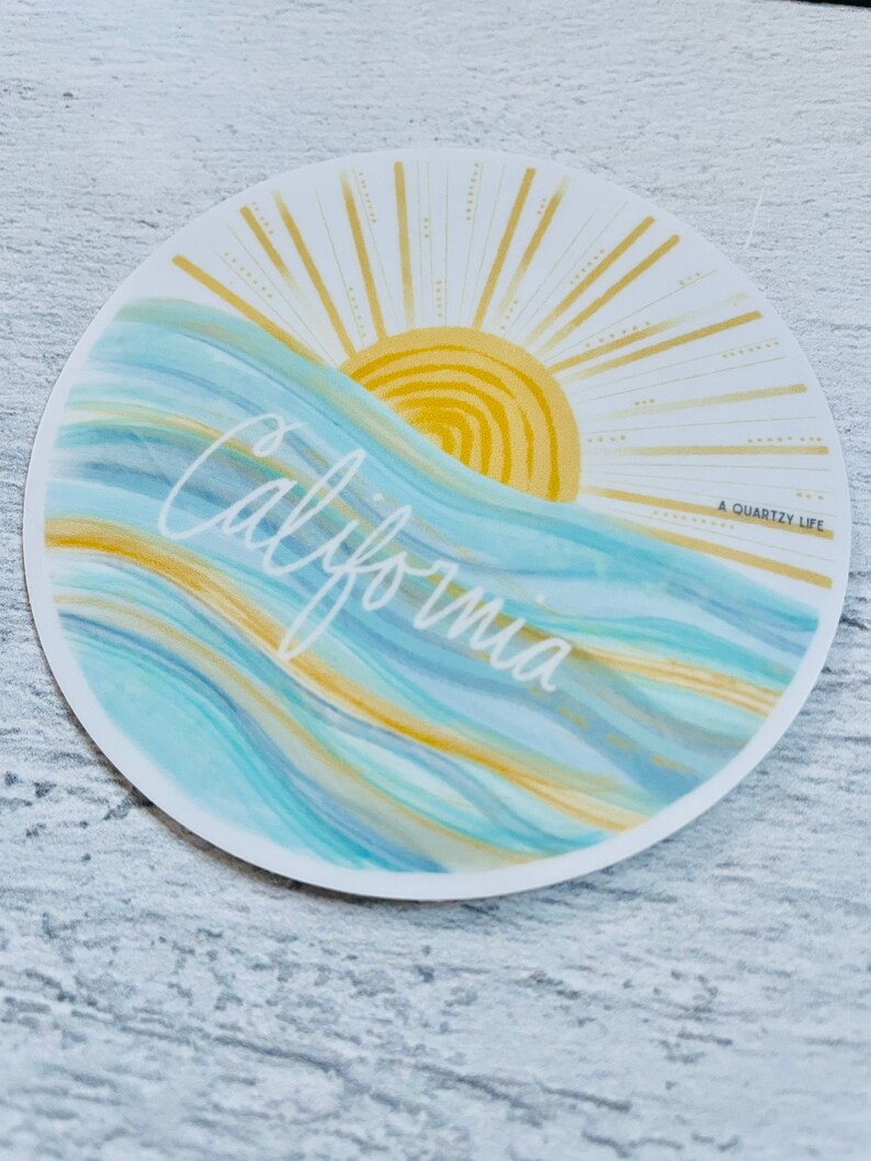 May include: A round sticker with a blue and yellow watercolor design of waves and a sun with rays. The text "California" is written in white cursive script and the text "A Quartzy Life" is written in small black letters in the bottom right corner.