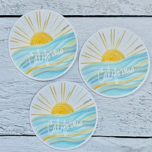 May include: Three round stickers with a sunset over the ocean and the word "California" written in white cursive. The stickers have a gold glitter background and a blue and white watercolor ocean.