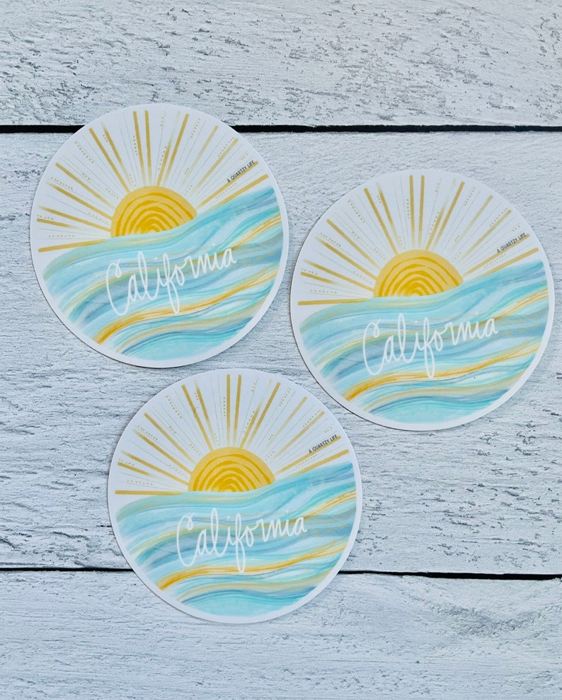 May include: Three round stickers with a sunset over the ocean and the word "California" written in white cursive. The stickers have a gold glitter background and a blue and white watercolor ocean.