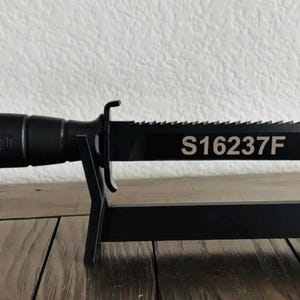 May include: A black knife with a serrated blade and the text "S16237F" on the blade. The knife is displayed on a black stand. The handle is cylindrical and textured.