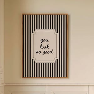 Pode incluir: Um interior de casa de banho com uma obra de arte emoldurada com riscas verticais a preto e branco e o texto "you look so good". Um pequeno banco de madeira suporta uma toalha branca enrolada e uma garrafa castanha. O chão tem um padrão xadrez a preto e branco.