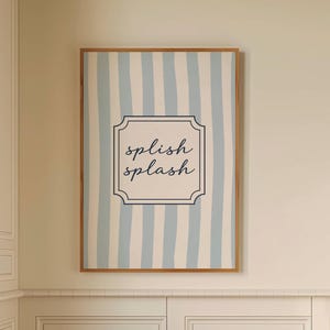Puede incluir: Un interior de baño con un marco que muestra rayas verticales azules y blancas y las palabras "splish splash". Un pequeño taburete de madera sostiene una toalla blanca enrollada y una botella marrón. La habitación tiene un suelo a cuadros blanco y negro.