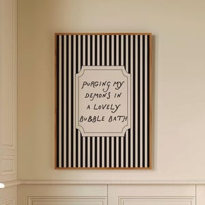 Puede incluir: Interior de baño con una obra de arte enmarcada con rayas verticales en blanco y negro y el texto "Purging my demons in a lovely bubble bath". Un taburete de madera sostiene una toalla y jabón. El suelo tiene un patrón de tablero de ajedrez en blanco y negro.