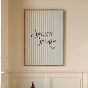 Puede incluir: Interior de baño con una obra de arte enmarcada que muestra "Splish Splash" en escritura cursiva sobre un fondo a rayas azul claro y blanco. Un taburete de madera sostiene una toalla blanca y una botella marrón. El suelo tiene un patrón de cuadros blancos y negros.