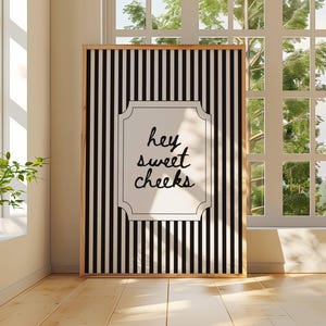 Puede incluir: Un cuadro enmarcado con un fondo a rayas blancas y negras y las palabras "hey sweet cheeks" en un marco decorativo. El cuadro está en una habitación con una ventana y plantas.