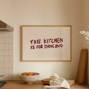 May include: A kitchen scene with a wooden countertop, a white sink, and a framed print that reads "THIS KITCHEN IS FOR DANCING." A bowl of oranges and a pan sit on the stovetop. A vase of white flowers is in the background.
