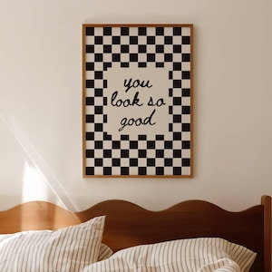 Puede incluir: Un cuadro enmarcado con un borde de patrón de tablero de ajedrez en blanco y negro. El centro del cuadro tiene las palabras "you look so good" en escritura negra. El cuadro está colgado sobre una cama con ropa de cama a rayas.