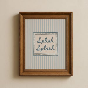 Puede incluir: Impresión enmarcada con un fondo a rayas azul claro y blanco. La impresión presenta las palabras "Splish Splash" en azul, escritas a mano dentro de un marco cuadrado. El marco es de color marrón cálido.