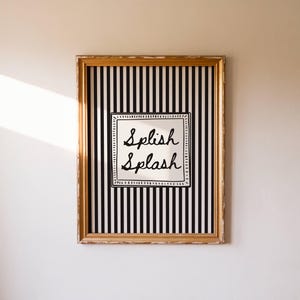 Könnte beinhalten: Ein gerahmtes Kunstwerk mit einem goldfarbenen Rahmen. Das Kunstwerk zeigt einen schwarz-weiß gestreiften Hintergrund mit einem weißen Quadrat in der Mitte. Die Worte "Splish Splash" sind in schwarzer Schrift innerhalb des weißen Quadrats geschrieben.