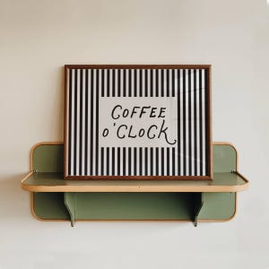 以下が含まれることがあります： 「COFFEE o' CLOCK」の文字が黒い筆記体で書かれたフレーム入りのプリント。白黒ストライプの背景に配置されています。プリントは、白い壁に取り付けられた緑と木目調の棚の上に置かれています。キッチンやダイニングエリア用の装飾品。