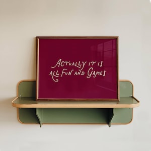 May include: A framed print with the words "ACTUALLY IT IS ALL FUN AND GAMES" in white script on a burgundy background. The frame is brown, and the print rests on a green and tan shelf against a white wall.