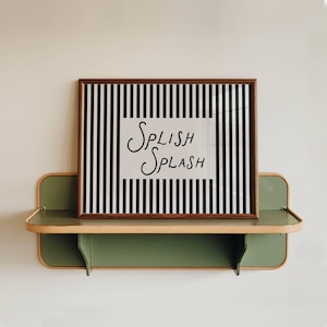 Puede incluir: Una obra de arte enmarcada con las palabras "Splish Splash" en escritura elegante. La obra presenta un fondo a rayas blancas y negras y un marco marrón. Descansa sobre una estantería verde y de madera, montada en una pared blanca.