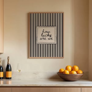 May include: A framed artwork with black and white vertical stripes and the text "how lucky are we" hangs above a countertop. Two champagne bottles, a wine glass, and a wooden bowl of oranges are on the counter.