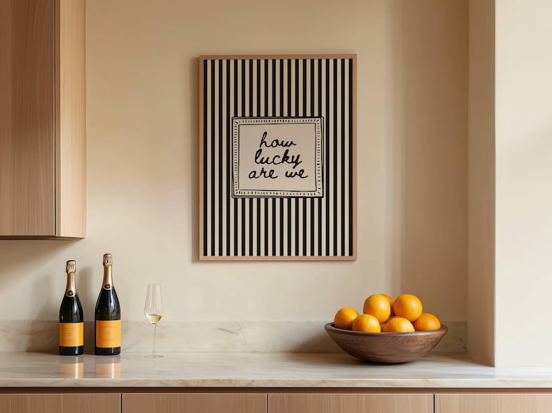 May include: A framed artwork with black and white vertical stripes and the text "how lucky are we" hangs above a countertop. Two champagne bottles, a wine glass, and a wooden bowl of oranges are on the counter.