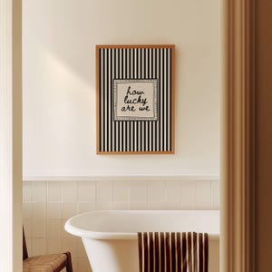 May include: A framed wall art piece with a black and white striped background and the text "how lucky are we" in a square frame. The artwork is displayed above a white bathtub in a bathroom setting.