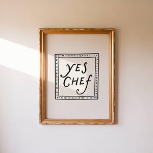 Puede incluir: Una impresión enmarcada con las palabras "YES CHEF" en escritura negra. El texto está dentro de un cuadrado con bordes negros y un borde punteado. El marco dorado y rectangular cuelga de una pared blanca.
