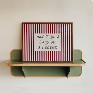 Può includere: Una stampa incorniciata con bordo marrone e la scritta "DON'T BE A LADY BE A LEGEND" su sfondo bianco. La stampa presenta un motivo a righe rosse e bianche. Poggia su una mensola verde e beige.