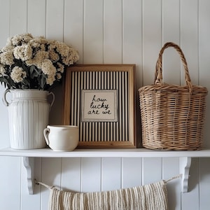 May include: A decorative shelf displays a white ceramic vase with flowers, a framed print with the text "how lucky are we", a small mug, and a woven basket. The print has a black and white striped background.