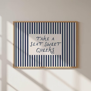 Puede incluir: Un estampado enmarcado con un fondo a rayas azul marino y blanco. El estampado presenta el texto "Take a seat sweet cheeks" en un estilo manuscrito. El marco es de color marrón claro.