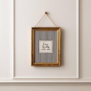 May include: A framed art piece with a gold-colored frame hanging on a white wall. The artwork features a black and white striped background with a square in the center that reads "how lucky are we". The frame is suspended by a thin, tan rope.