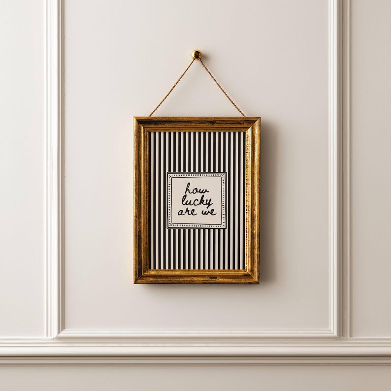 May include: A framed art piece with a gold-colored frame hanging on a white wall. The artwork features a black and white striped background with a square in the center that reads "how lucky are we". The frame is suspended by a thin, tan rope.