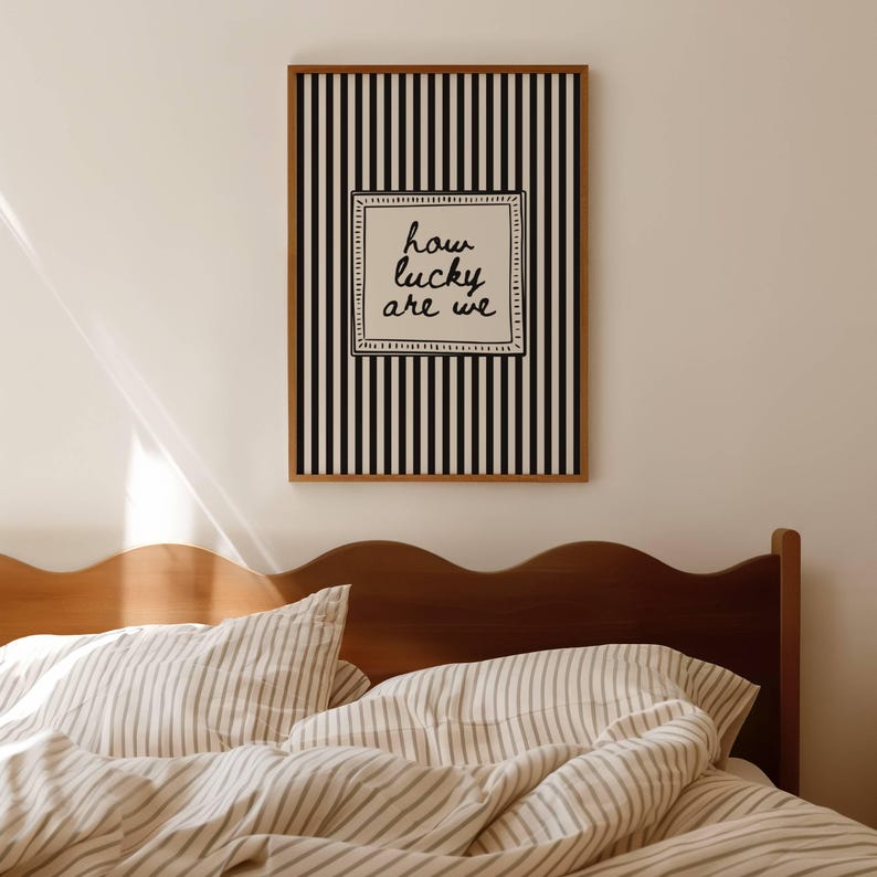 May include: A framed wall art print with a black and white striped background. The print features the phrase "how lucky are we" in black script within a white square. The frame is a warm brown color. The image is above a bed with striped bedding.
