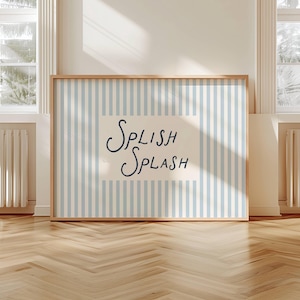 Puede incluir: Una obra de arte enmarcada con un fondo a rayas azul claro y blanco. La obra presenta las palabras "SPLISH SPLASH" en una fuente manuscrita. El marco es de madera clara y la obra se exhibe sobre un suelo de madera.