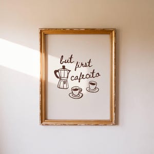 May include: A framed print with a gold-colored frame. The print features a brown illustration of a coffee maker, two coffee cups, and the words "but first cafecito" in a handwritten style. The background is white.