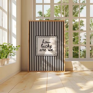 May include: A framed art print with a black and white striped background and the text "how lucky are we" in a white square. The print is displayed in a bright room with a window and plants.