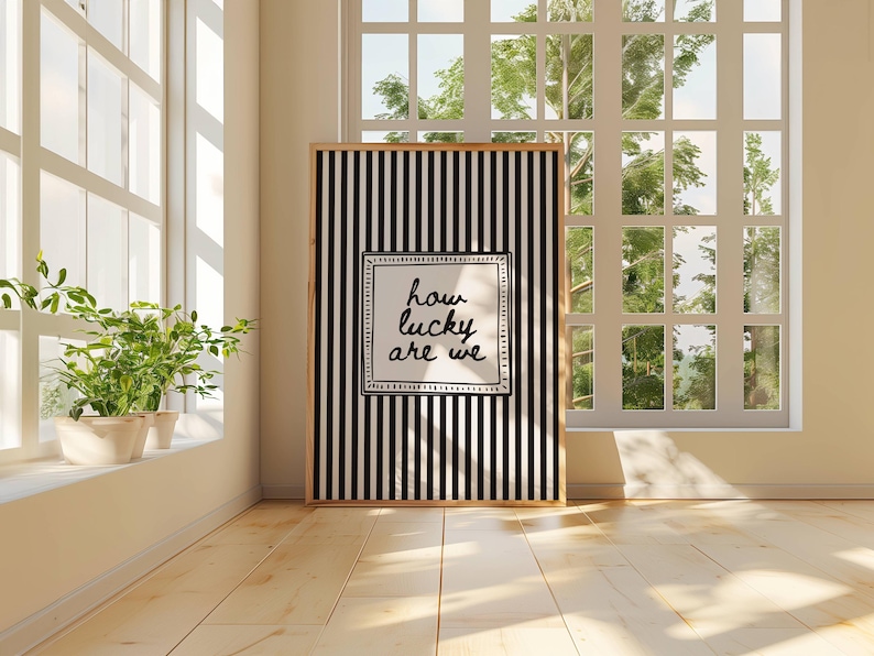May include: A framed art print with a black and white striped background and the text "how lucky are we" in a white square. The print is displayed in a bright room with a window and plants.