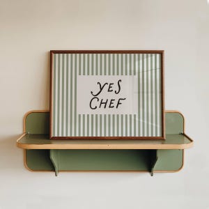 Puede incluir: Una impresión enmarcada con el texto "YES CHEF" se encuentra en un estante verde y de madera. La impresión tiene un fondo a rayas en blanco y verde claro. El estante está montado en una pared blanca.