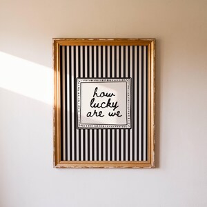May include: A framed artwork with a gold-colored frame. The artwork features a black and white striped background with a white square in the center. The text "how lucky are we" is written in black script within the white square.