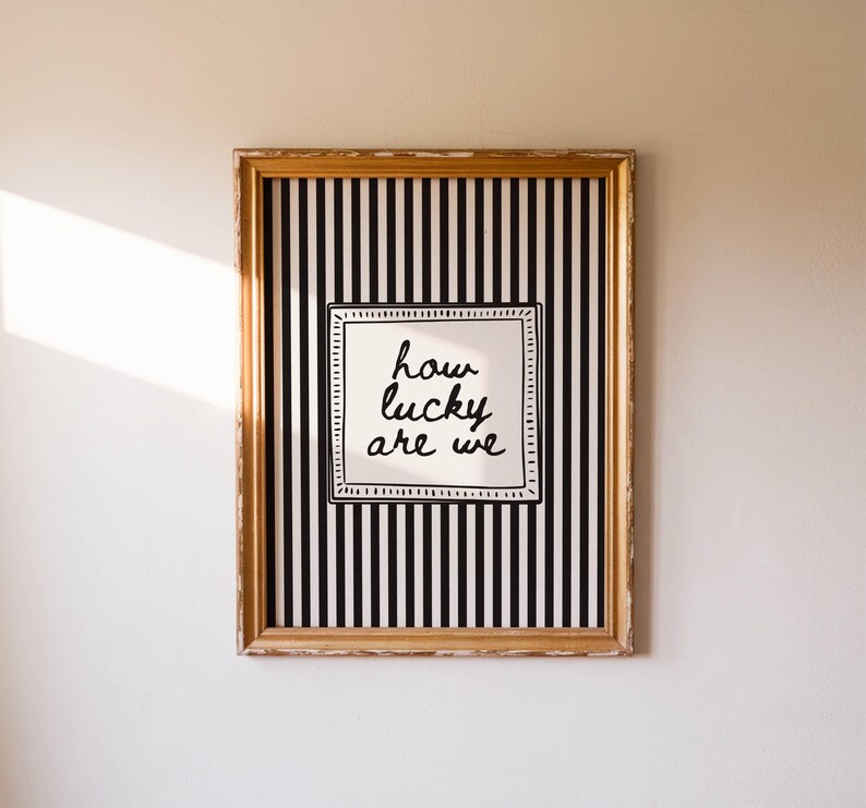 May include: A framed artwork with a gold-colored frame. The artwork features a black and white striped background with a white square in the center. The text "how lucky are we" is written in black script within the white square.