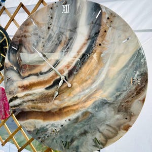 May include: Large, round wall clock with a marbled design in shades of gray, brown, and cream. The clock face features silver Roman numerals and hands. The clock is hanging from a metal hook.