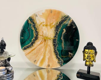 Puede incluir: Reloj de pared redondo con un diseño de resina verde, dorada y beige. La esfera del reloj presenta la palabra "Family" en letras doradas. El reloj tiene manecillas doradas y un borde negro. El reloj se muestra sobre una superficie reflectante.