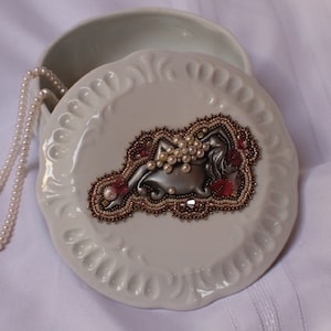 May include: A white ceramic jewelry box with a decorative lid featuring a beaded design of a reclining figure. The design incorporates pearls, red accents, and metallic elements. A pearl necklace rests beside the box.