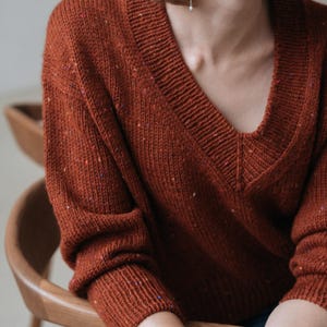May include: A terracotta-coloured V-neck sweater with a textured knit. The sweater has a relaxed fit and long sleeves. The fabric has flecks of colour throughout. The person is wearing a silver bracelet and pearl earrings. The sweater is a cosy, casual garment.