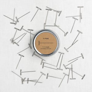 Può includere: Spilli a T in acciaio inossidabile sparsi attorno a una scatola rotonda. La scatola ha un'etichetta marrone con la scritta "T-PINS" e "1 1/2 in length, 40 stainless steel pins". Gli spilli sono lunghi circa 3,8 cm.