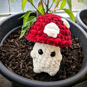 May include: A handmade, plush mushroom figure with a red cap and white spots, and a white stem. The mushroom is planted in a black pot with soil, and a plant with green leaves is growing out of the top. The background is blurred.