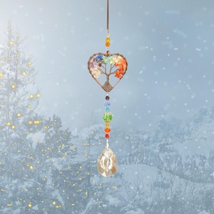 Puede incluir: Un atrapasol de cristal en forma de corazón con un diseño de árbol de la vida y cristales de colores arcoíris que cuelgan de él. El atrapasol está colgado frente a un fondo nevado.