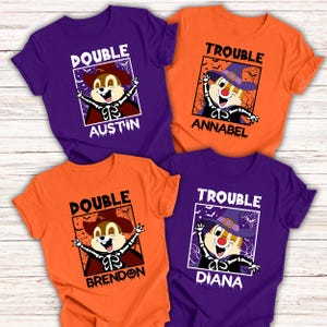 Può includere: Quattro magliette a maniche corte viola e arancioni. Ogni maglietta presenta un personaggio di scoiattolo dei cartoni animati in costume di Halloween con le parole "DOUBLE" o "TROUBLE" sopra il personaggio e un nome sotto.