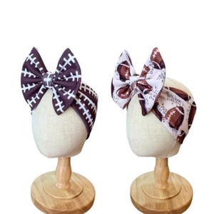 May include: Two headbands displayed on stands. The left headband is dark brown with a white football lace pattern and a large bow. The right headband is white with a brown football print and a large bow. Both feature a wide, fabric band design.