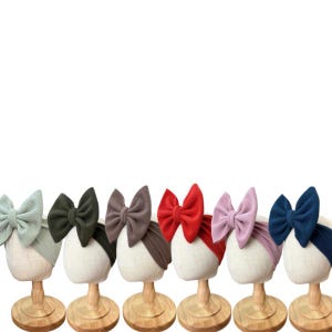 May include: A set of six ribbed fabric headbands, each featuring a large, prominent bow. The headbands are displayed in a row on individual stands and come in light green, dark green, brown, red, light purple, and dark blue color variations.
