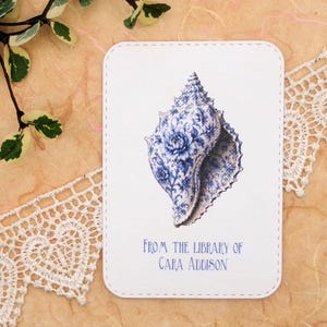 May include: A white card with a blue and white seashell illustration. The seashell has a floral design. The text "FROM THE LIBRARY OF CARA ADDISON" is printed in blue at the bottom of the card. The card has rounded corners and a stitched border.