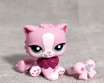 Puede incluir: Una figura de gato rosa con una raya blanca en la cara, ojos verdes y un collar rosa oscuro. Un anillo rosa en forma de lazo con una gema azul está al lado del gato. El gato tiene patas rosas con almohadillas blancas.