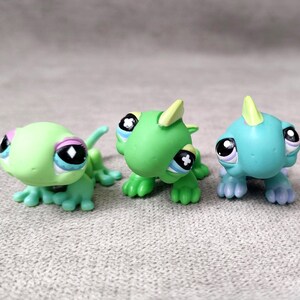 May include: Three colorful lizard figurines in shades of green and blue. Each figurine has large, expressive eyes with black pupils and white highlights. The lizards have small, spiky crests and are posed on a neutral surface.